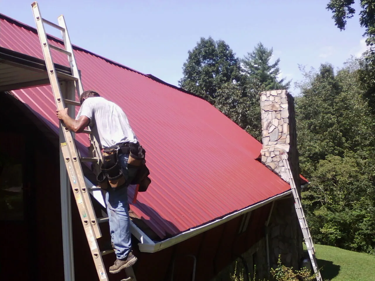 Roofer on a ladder beside a red metal roof and stone chimney — Soffit & Fascia Repair by Roofing Brothers in Gillette